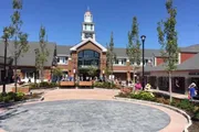 The image shows a sunny day at a well-kept outdoor shopping center featuring a building with a prominent clock tower, surrounded by shoppers enjoying the day.