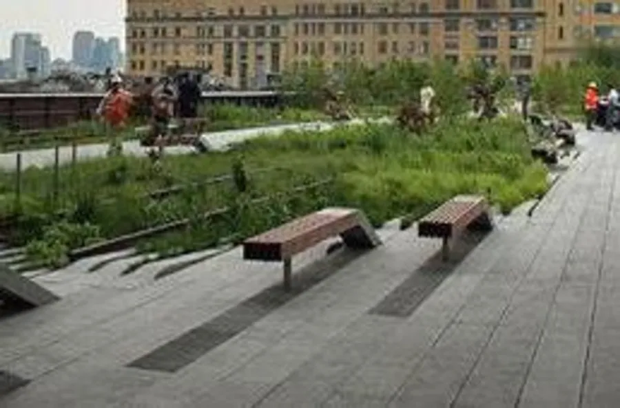 The image shows a modern urban park with wooden benches and lush greenery, against a backdrop of city buildings, with people walking along the path.