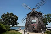 The image features a traditional windmill by the water with people walking nearby, set against a clear blue sky.