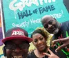 Three people are posing with smiles and hand signs in front of a colorful wall with graffiti that reads The Graffiti Hall of Fame
