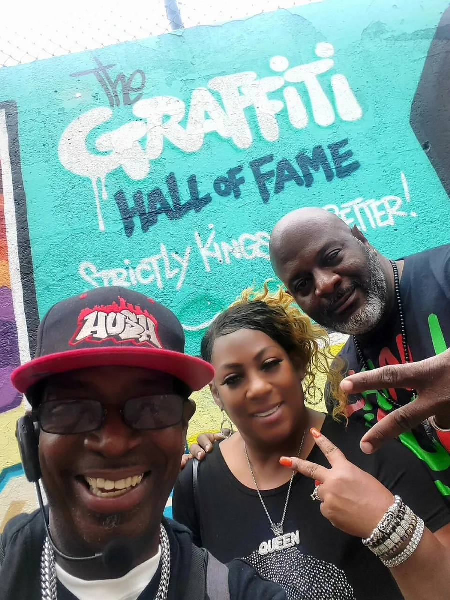 Three people are posing with smiles and hand signs in front of a colorful wall with graffiti that reads The Graffiti Hall of Fame.