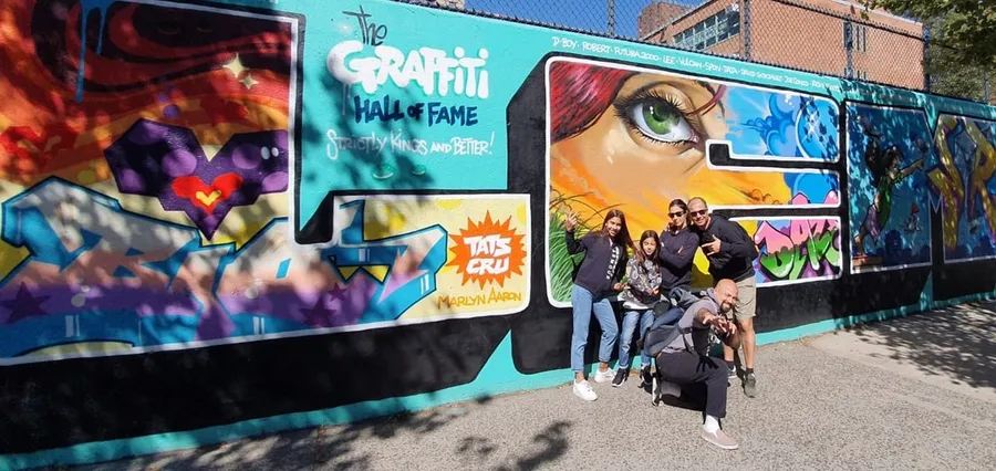 A group of people is posing for a photo in front of a vibrant graffiti wall that includes the text The Graffiti Hall of Fame, colorful artwork, and a large depiction of an eye.