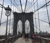 Pedestrians stroll along the wooden walkway of the iconic Brooklyn Bridge flanked by its distinctive web of cables and towering Gothic-style stone pillars against a backdrop of a partly cloudy sky