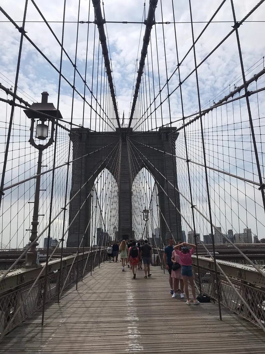 Pedestrians stroll along the wooden walkway of the iconic Brooklyn Bridge, flanked by its distinctive web of cables and towering Gothic-style stone pillars, against a backdrop of a partly cloudy sky.