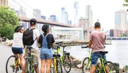 A group of cyclists is pausing to enjoy the view of the city skyline and a bridge from a riverside promenade.