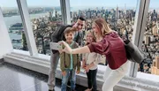 A family is taking a selfie with a smartphone inside a high-rise building, overlooking a panoramic cityscape.