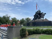 A military band in red uniforms stands in formation beside the iconic United States Marine Corps War Memorial statue, against a backdrop of trees, buildings, and a waving American flag.
