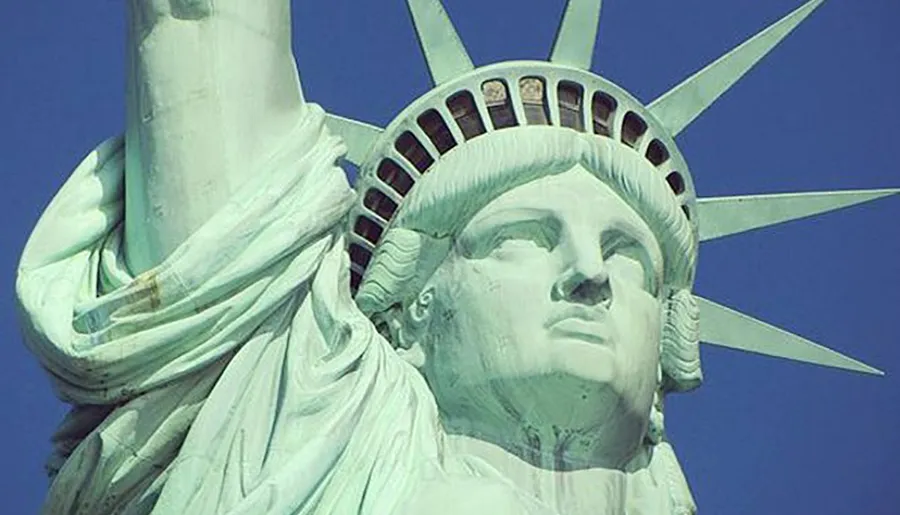 The image is a close-up of the face and crown of the Statue of Liberty against a clear blue sky.