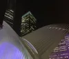 The image shows the illuminated wing-like structure of the Oculus at the World Trade Center transportation hub in New York City with skyscrapers towering in the background at night