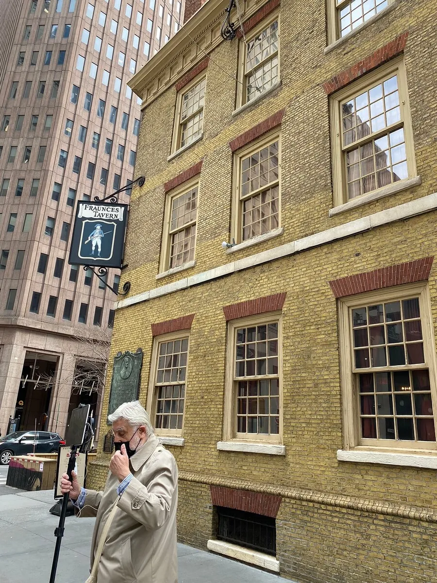 A person is looking at a mobile device in front of Fraunces Tavern with a historical building plaque, situated amid contrasting modern architecture.