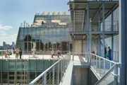 This image shows people enjoying a sunny day on a modern rooftop terrace with glass structures and a view of a city skyline.