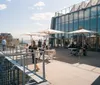 This image shows people enjoying a sunny day on a modern rooftop terrace with glass structures and a view of a city skyline