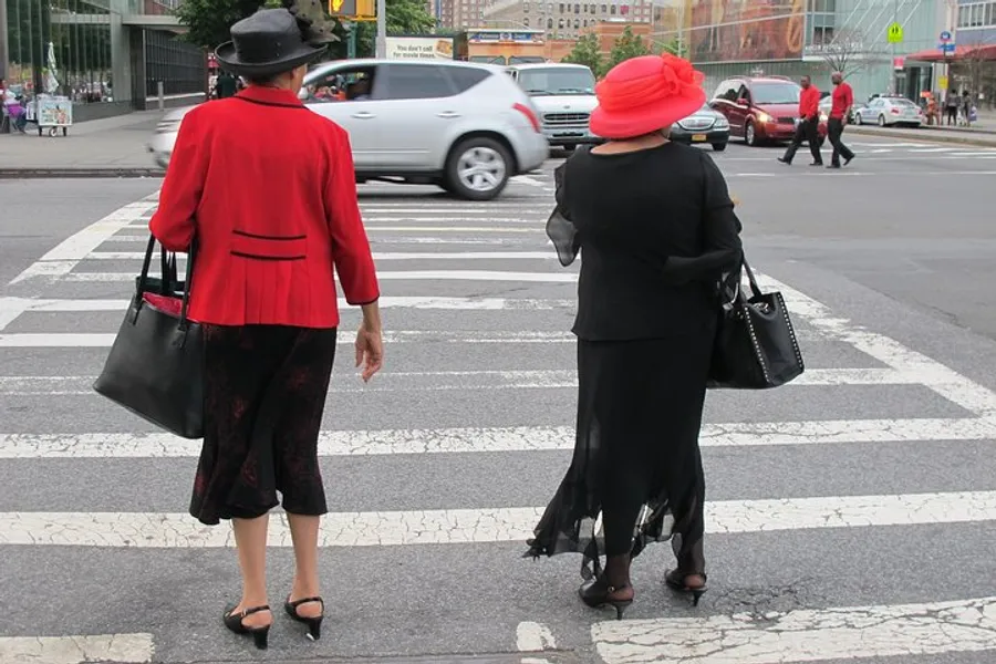 Two elegantly dressed individuals, one in a red outfit and the other in black, are standing at a pedestrian crossing in an urban setting.