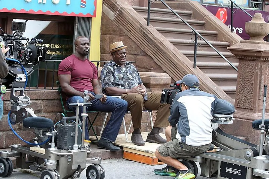 Two actors are seated on a stoop during a film or television shoot while a camera operator captures the scene.