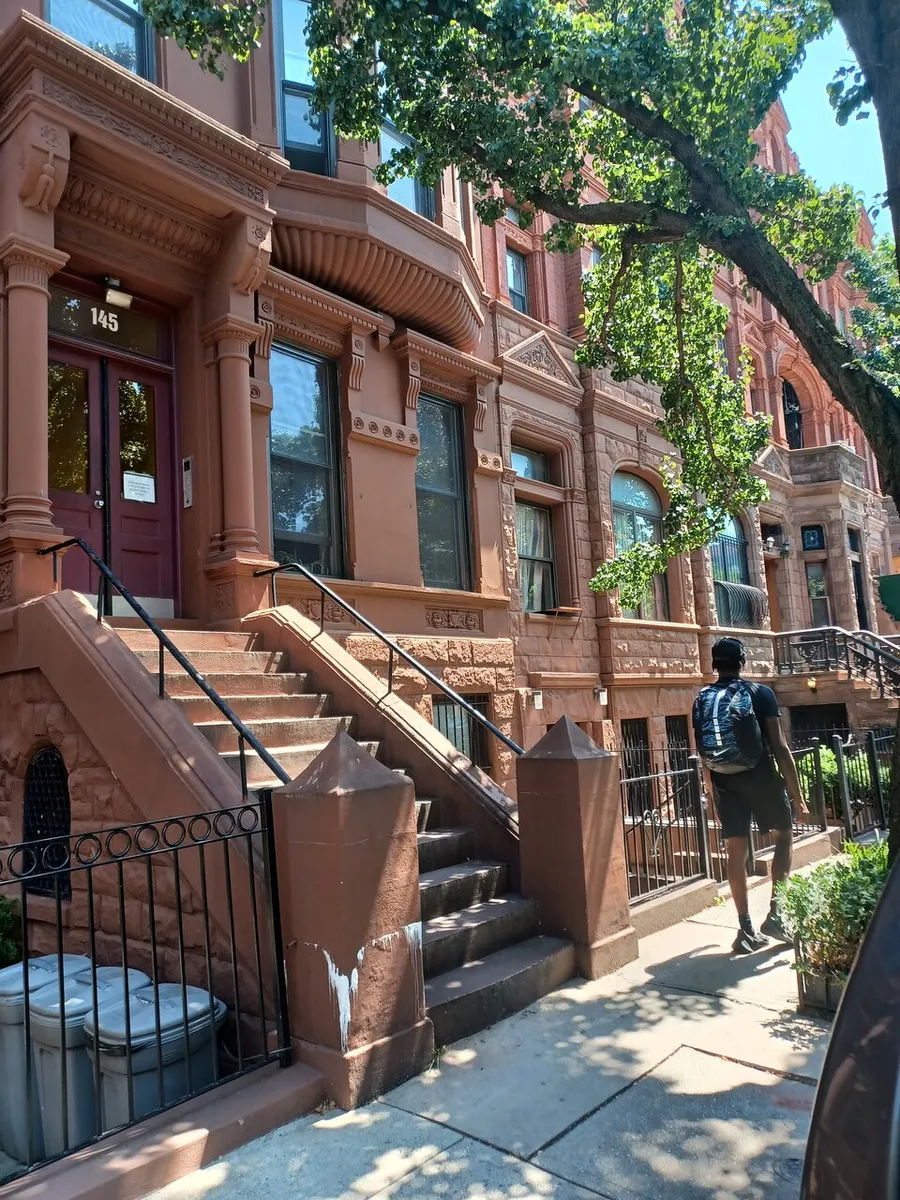 A person walks past the entrance to a brownstone building with ornate detailing and a stoop in a tree-lined urban street.