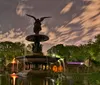 The image captures a nighttime view of the illuminated Bethesda Fountain in Central Park New York City with a reflective pool in the foreground and ambient city lights peering through the trees in the background