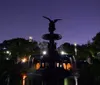 The image captures a nighttime view of the illuminated Bethesda Fountain in Central Park New York City with a reflective pool in the foreground and ambient city lights peering through the trees in the background