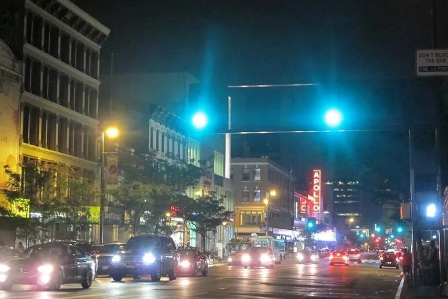 The image shows a bustling urban street at night, highlighted by the illuminated signage of the Apollo Theater, with cars on the road and a dynamic cityscape backdrop.