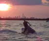 Two individuals are riding jet skis on a body of water at sunset with a city skyline visible in the background