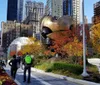 The image shows pedestrians walking through an urban park with colorful autumn foliage with a large abstract bronze sculpture in the background set against a backdrop of towering buildings