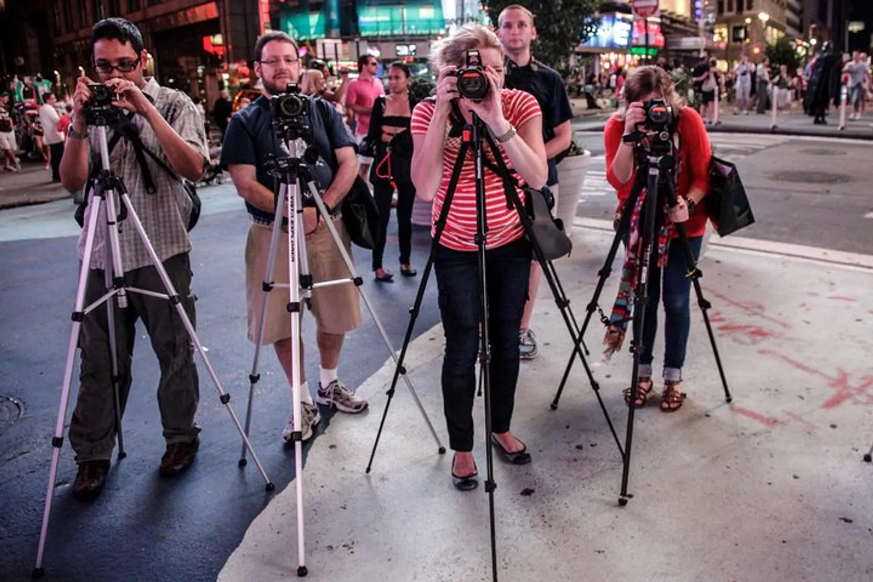 A group of photographers with their cameras mounted on tripods are taking pictures in a bustling urban setting at night.