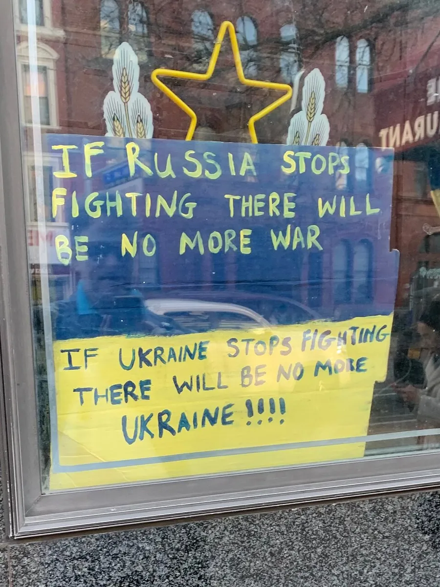 The image depicts a handwritten sign with a statement about the cessation of fighting by Russia leading to no more war, and a contrasting statement about Ukraine, likely expressing a political viewpoint on an ongoing conflict.