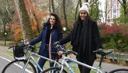 Two smiling women are standing with their bicycles in a park with autumn foliage in the background.