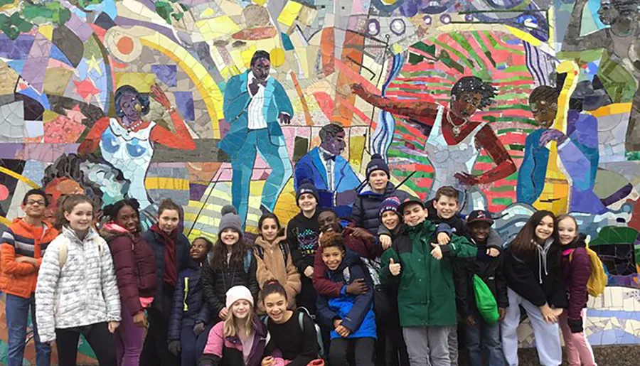 A group of smiling children poses in front of a colorful mosaic wall featuring artistic depictions of people.