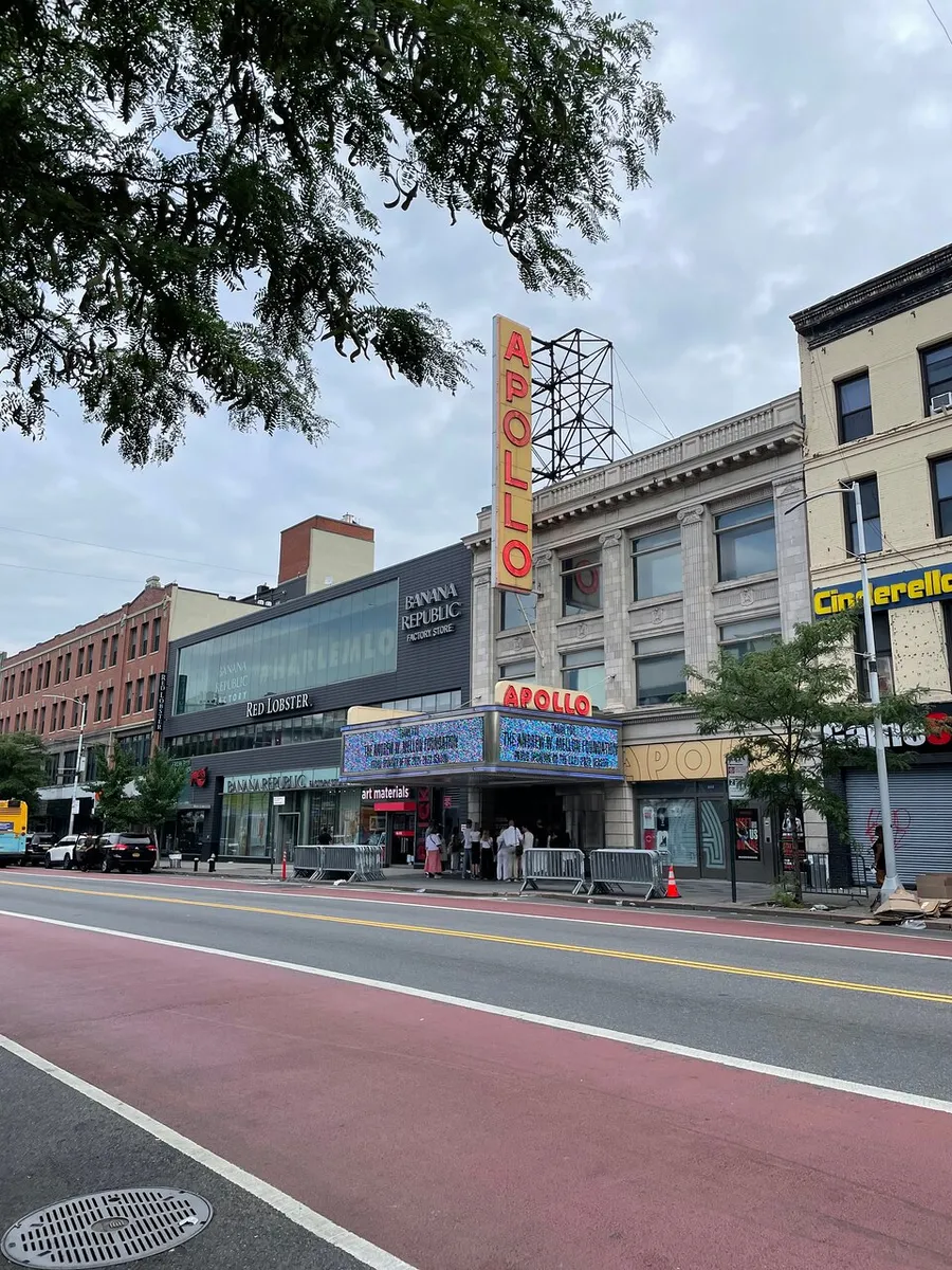 The image shows the iconic Apollo Theater with its notable neon sign, located on a city street with pedestrians and various storefronts, under an overcast sky.
