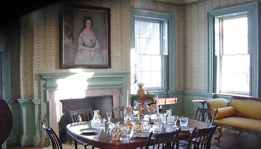 The image shows an elegantly set historical dining room with a large oval table, glassware, a fireplace, traditional furniture, a portrait painting on the wall, and two windows allowing in natural light.