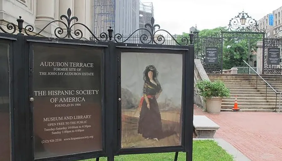 The image shows a gated entrance to a cultural institution, with informational signage about The Hispanic Society of America and a portrait on display.