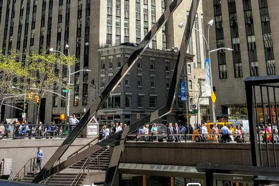 This image captures a bustling city scene with a crowd of people on a sidewalk, modern buildings, a subway entrance, and a distinctive sculptural metallic structure under a clear blue sky.