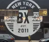 The image shows the exterior of a building painted black with a large white circular logo that reads New York City BX Bronx Brewery EST 2011 indicating the establishment is a brewery in the Bronx New York City established in 2011 with a person wearing a helmet standing in front of it