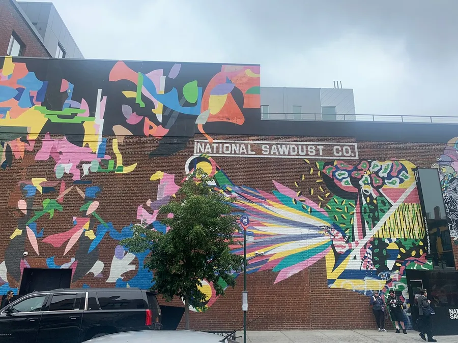The image showcases a vibrant, abstract mural on the exterior of a brick building with a sign that reads National Sawdust Co., and several people are present near the entrance.