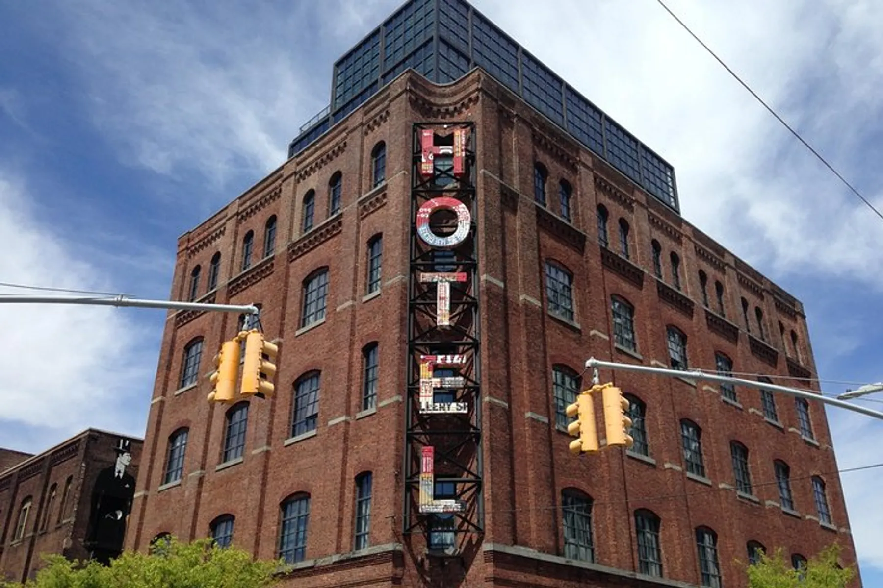 The image shows a tall brick building with a unique triangular shape featuring a fire escape and large letter signage on its side, set against a blue sky with scattered clouds, interspersed with traffic lights from an urban street perspective.