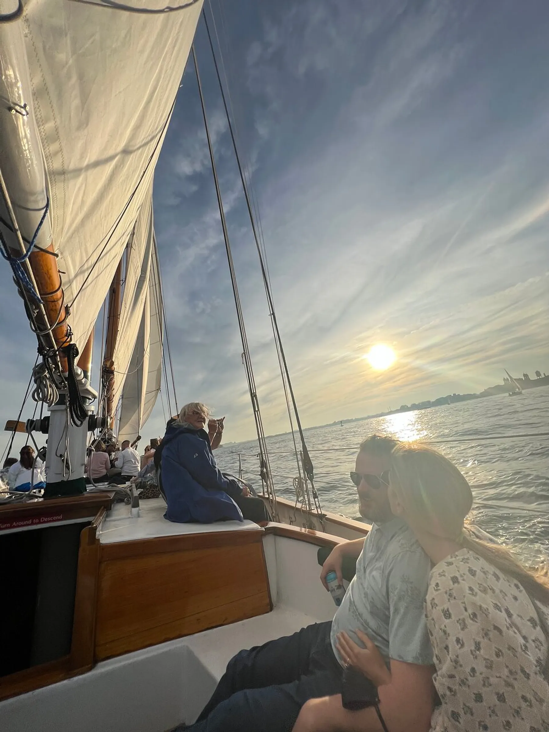 People are enjoying a serene sailboat ride at sunset, with the sails hoisted against the backdrop of a calm sea and the setting sun.