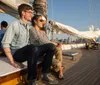 A couple is sitting on the deck of a sailboat smiling and looking out towards the city skyline at sunset