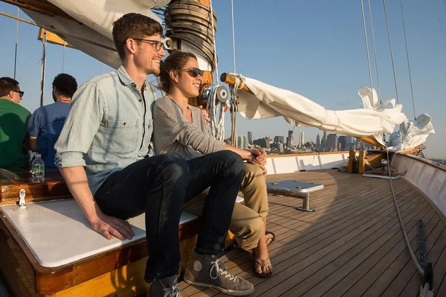 A couple is sitting on the deck of a sailboat, smiling and looking out towards the city skyline at sunset.