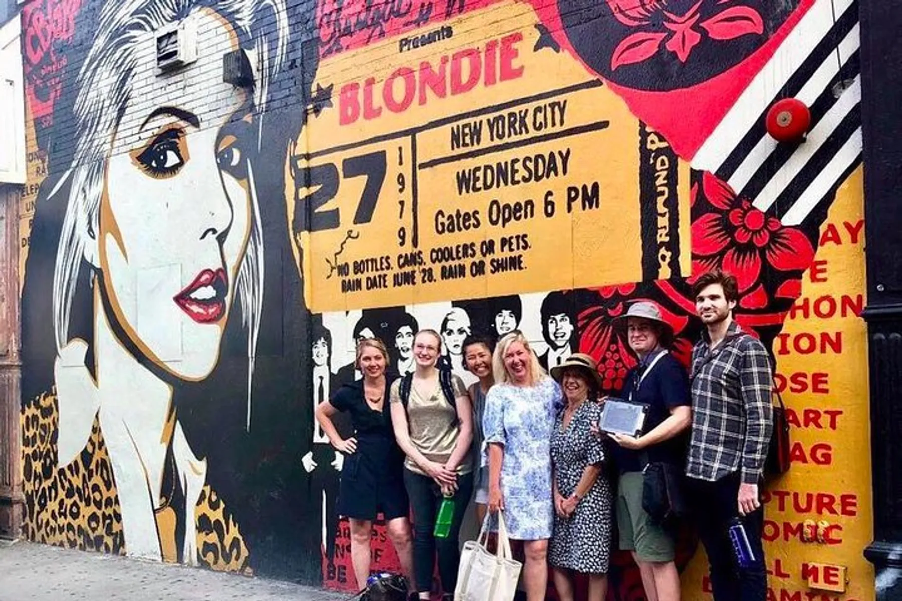A group of people is smiling for the camera in front of a vibrant street mural advertising a Blondie concert in New York City.