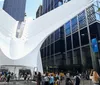 The image shows a bustling urban scene with people walking in front of the distinctive modern white ribbed structure of the Oculus in New York City