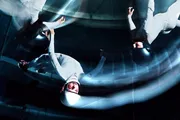 The image depicts individuals in white suits and helmets seemingly floating or flying within a wind tunnel, captured with a motion blur effect.