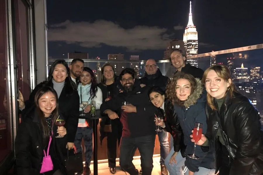 A group of people are posing for a photo at night with drinks in their hands on a rooftop overlooking a city skyline that includes the Empire State Building.
