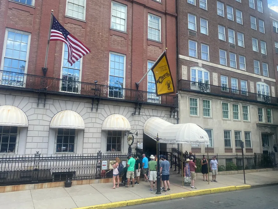 A group of people is queuing outside a building flying the American flag and a yellow banner, with a striped awning over the entrance.