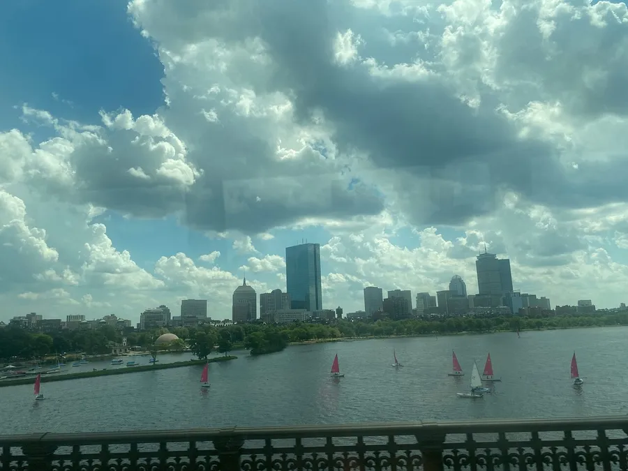 The image shows a view of a city skyline across a body of water with sailboats and a cloudy blue sky, as seen through a window with reflections.