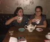Two individuals are smiling and holding teacups in a cozy setting with vintage wallpaper and a teapot on the table suggesting a casual and friendly tea time