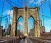 The image showcases pedestrians walking along the Brooklyn Bridge pedestrian pathway with the distinctive gothic arches of the bridge framing a view of the Manhattan skyline