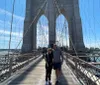The image showcases pedestrians walking along the Brooklyn Bridge pedestrian pathway with the distinctive gothic arches of the bridge framing a view of the Manhattan skyline