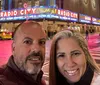 A smiling man and woman are taking a selfie in front of the brightly lit marquee of Radio City Music Hall at night