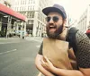 A cheerful man with a beard and sunglasses is walking on a city street holding a paper bag seemingly in mid-laughter or conversation