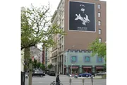 A person riding a bicycle on an urban street with a large Dior Homme advertisement featuring a shark on the side of a building.
